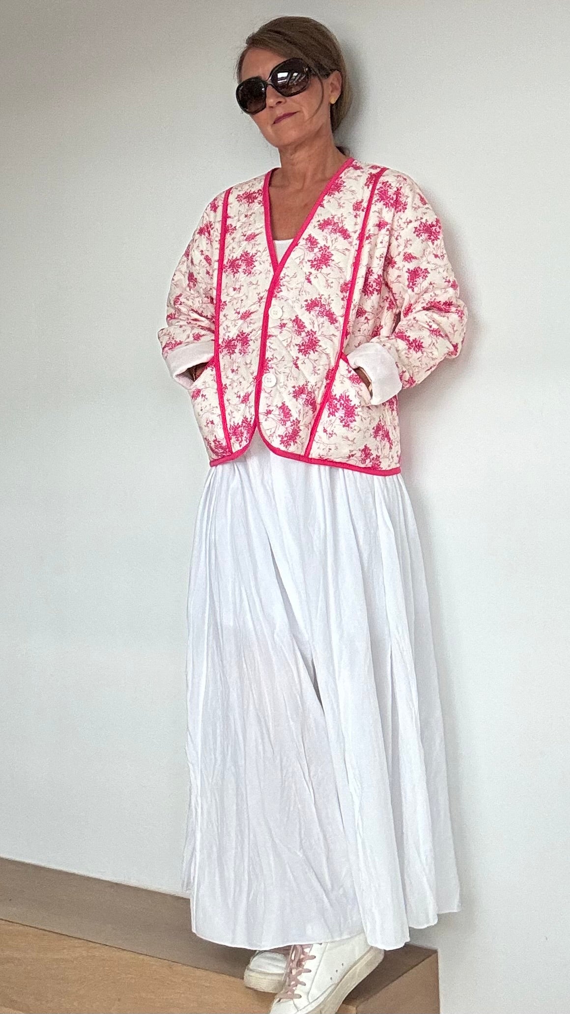 Steppjacke aus 100% Baumwolle in strahlendem Weiß mit pinkem Blumenmuster. Die Jacke kann offen oder mit eleganten Knöpfen geschlossen getragen werden. Mit einem oversized Schnitt und einer Rückenlänge von 56 cm passt sie perfekt über weite Kleider oder Röcke. Besondere Details sind aufgesetzte Taschen und feine Ziernähte. Ideal für kühlere Tage und bietet hohen Tragekomfort.
