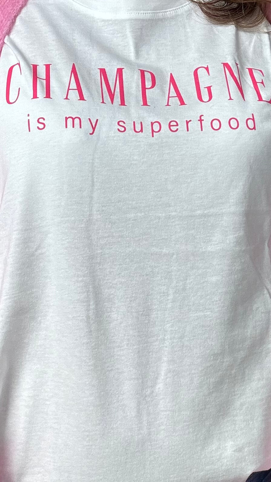 Cremeweißes T-Shirt mit einem lockeren Halsausschnitt und kurzen Ärmeln, auf der Brust steht in Pink geschreiben: Champagne is my superfood. Details findest du in unserer Produktbeschreibung.