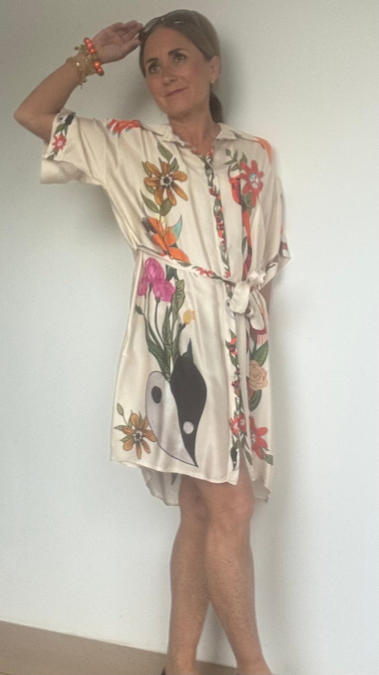 Blusenkleid aus 100 % Viskose mit ecrufarbenem Grundton und floralem Muster. Länge ca. 91 cm, locker geschnitten, mit Knopfleiste und Bindegürtel in der Taille. Auf der Rückseite des Kleides ist eine Göttin auf einem Leopard abgebildet – ein auffälliges, mystisch inspiriertes Detail. Das Kleid wird einmal geschlossen als Kleid und einmal offen als Kaftan über einer Hose gezeigt