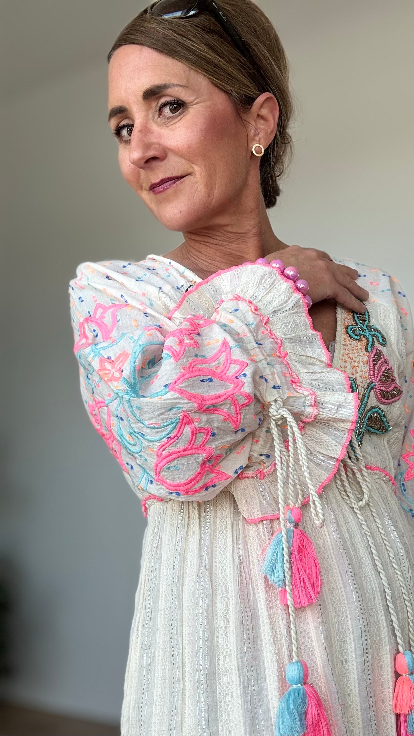 Maxi-Kleid „Boho Blossom“ in Ecru mit silbernen Fäden, besticktem V-Ausschnitt mit Troddelbändern, langen Ärmeln mit farbiger Stickerei in Pink und Hellblau, sowie Perlen- und Paillettenverzierung. Gummizug unter der Brust, Achselweite 49 cm, Länge 135 cm. Ideal für Frauen ab 160 cm Körpergröße. 100 % Baumwolle mit Unterkleid.
