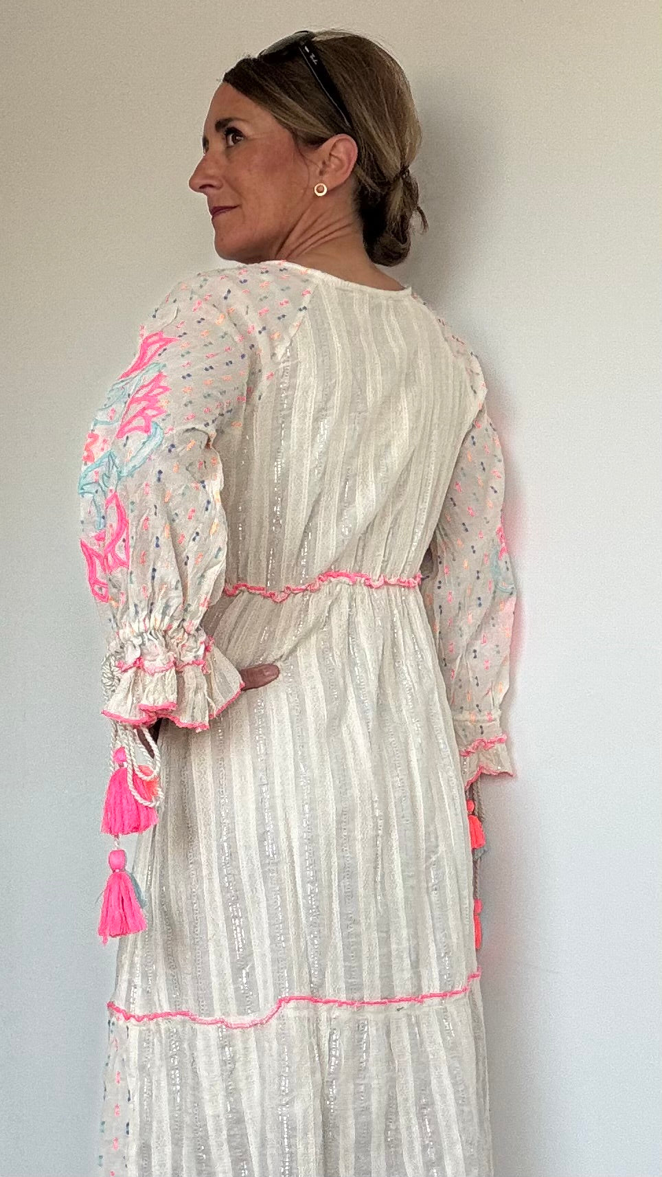 Maxi-Kleid „Boho Blossom“ in Ecru mit silbernen Fäden, besticktem V-Ausschnitt mit Troddelbändern, langen Ärmeln mit farbiger Stickerei in Pink und Hellblau, sowie Perlen- und Paillettenverzierung. Gummizug unter der Brust, Achselweite 49 cm, Länge 135 cm. Ideal für Frauen ab 160 cm Körpergröße. 100 % Baumwolle mit Unterkleid.