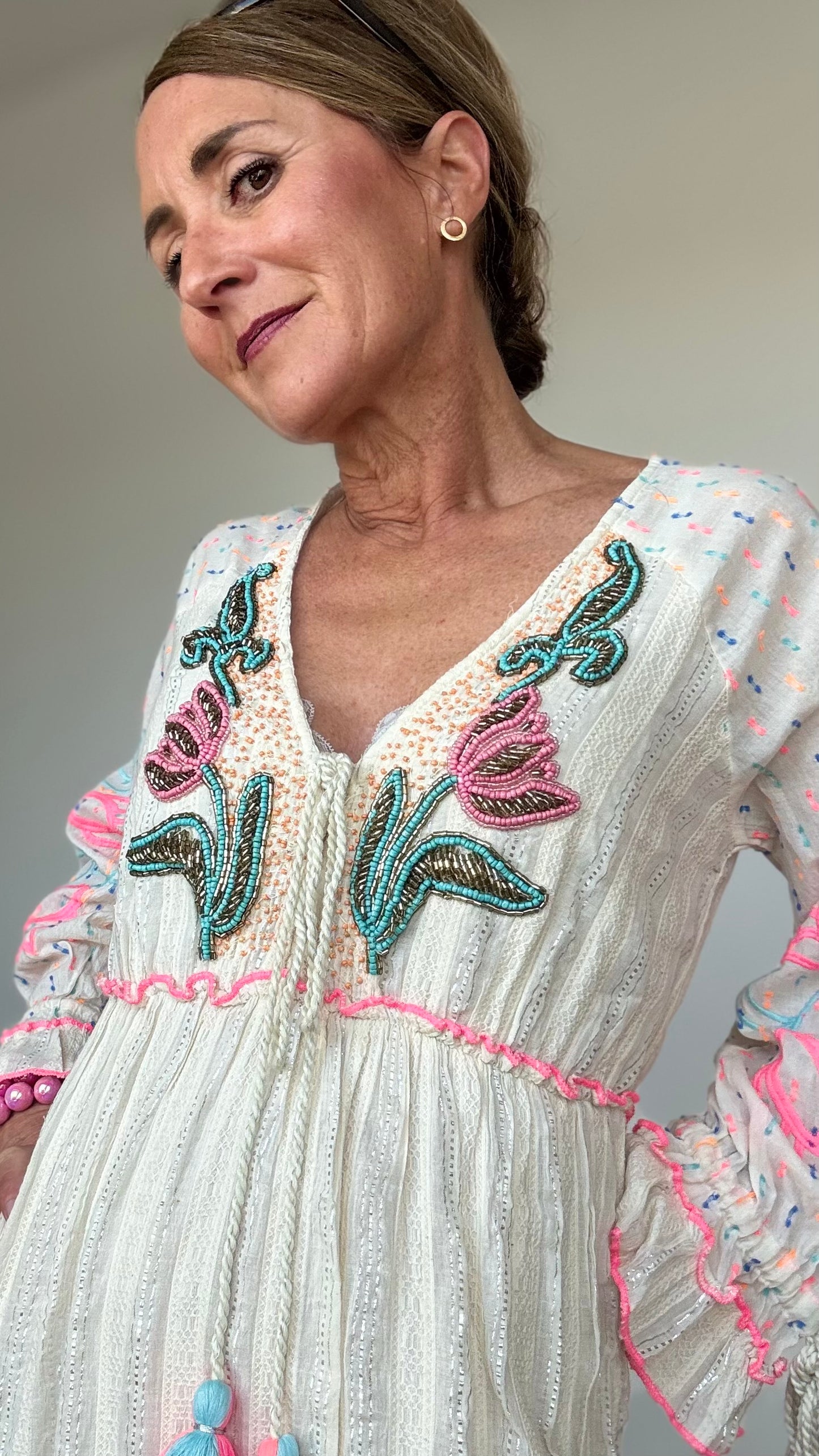 Maxi-Kleid „Boho Blossom“ in Ecru mit silbernen Fäden, besticktem V-Ausschnitt mit Troddelbändern, langen Ärmeln mit farbiger Stickerei in Pink und Hellblau, sowie Perlen- und Paillettenverzierung. Gummizug unter der Brust, Achselweite 49 cm, Länge 135 cm. Ideal für Frauen ab 160 cm Körpergröße. 100 % Baumwolle mit Unterkleid.