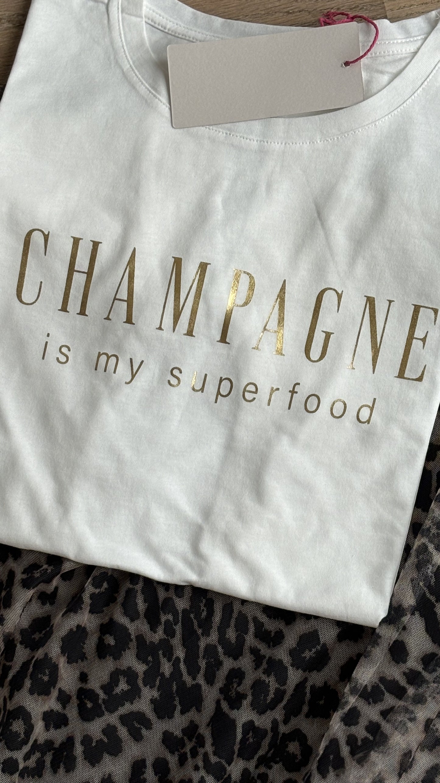 Cremeweißes T-Shirt mit einem lockeren Halsausschnitt und kurzen Ärmeln, auf der Brust steht in Pink geschreiben: Champagne is my superfood. Details findest du in unserer Produktbeschreibung.