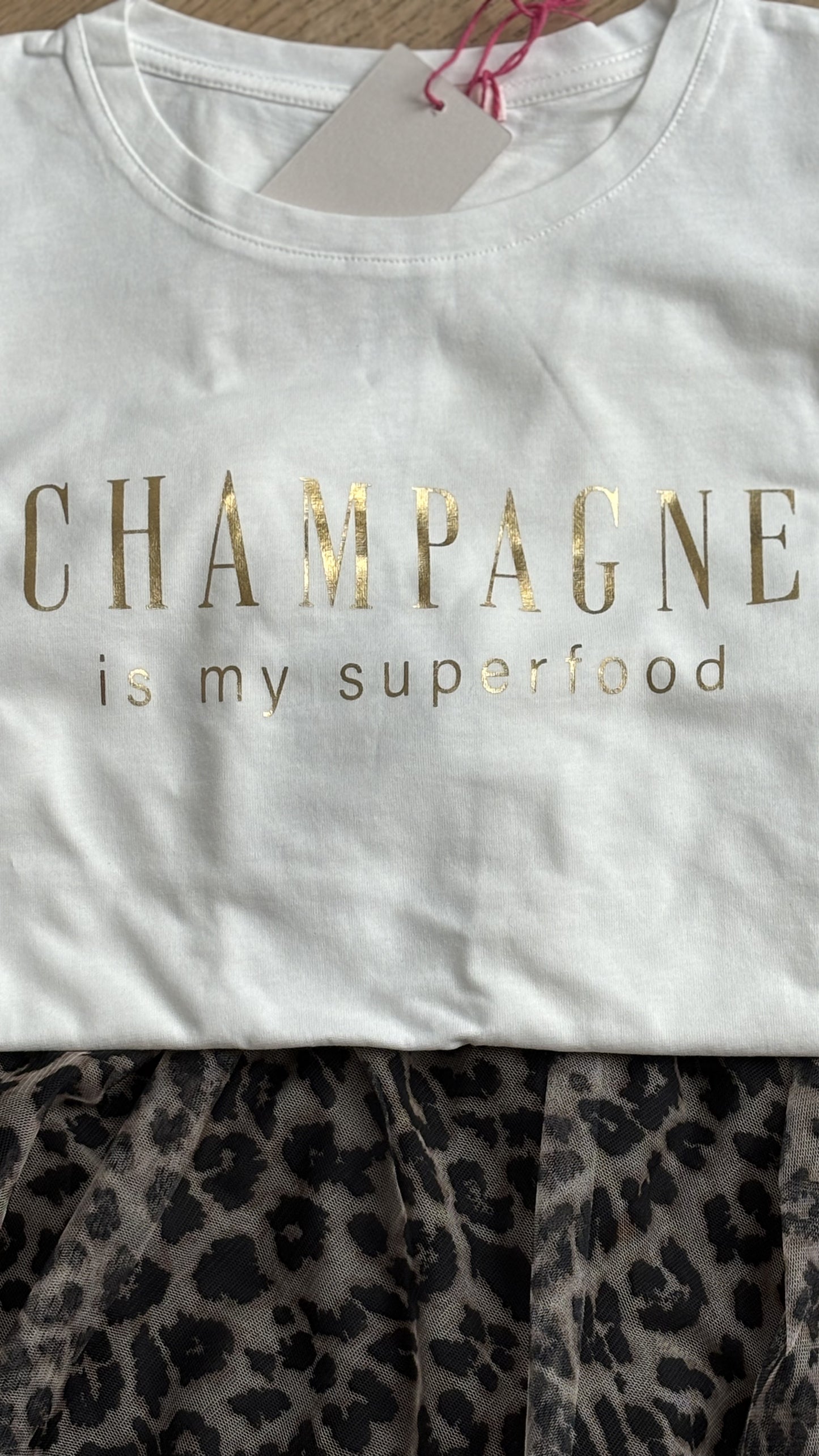 Cremeweißes T-Shirt mit einem lockeren Halsausschnitt und kurzen Ärmeln, auf der Brust steht in Pink geschreiben: Champagne is my superfood. Details findest du in unserer Produktbeschreibung.