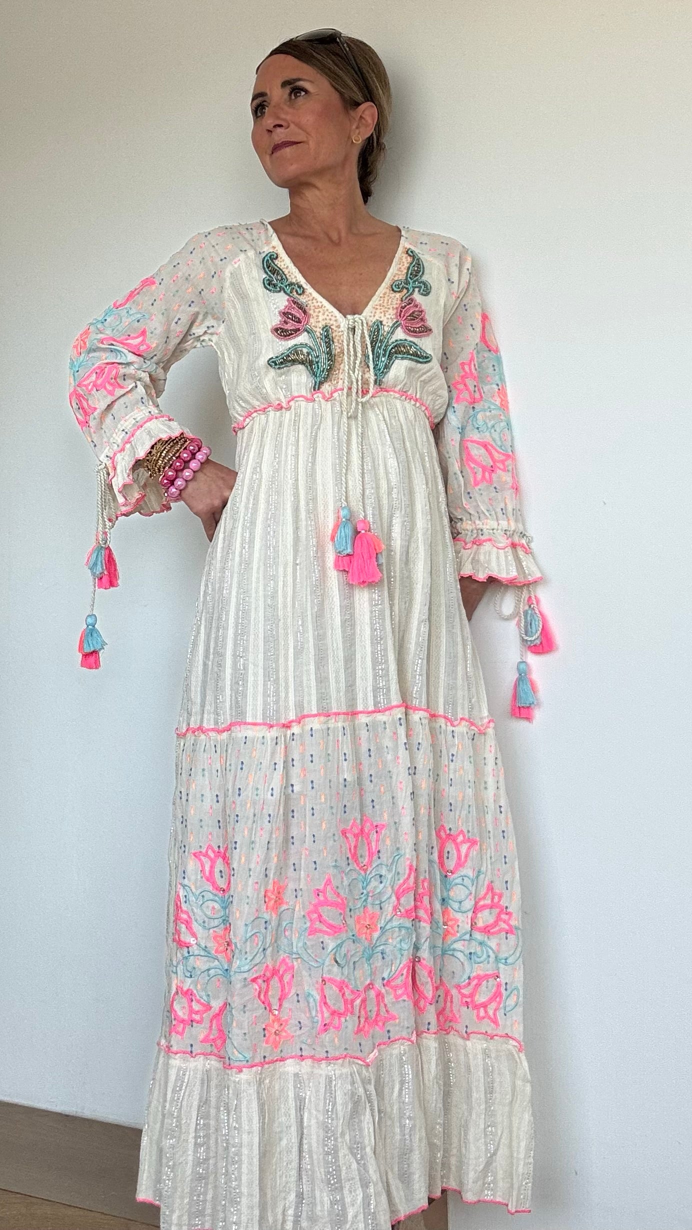 Maxi-Kleid „Boho Blossom“ in Ecru mit silbernen Fäden, besticktem V-Ausschnitt mit Troddelbändern, langen Ärmeln mit farbiger Stickerei in Pink und Hellblau, sowie Perlen- und Paillettenverzierung. Gummizug unter der Brust, Achselweite 49 cm, Länge 135 cm. Ideal für Frauen ab 160 cm Körpergröße. 100 % Baumwolle mit Unterkleid.