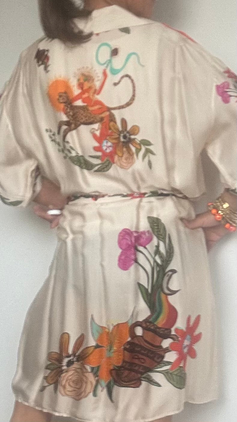 Blusenkleid aus 100 % Viskose mit ecrufarbenem Grundton und floralem Muster. Länge ca. 91 cm, locker geschnitten, mit Knopfleiste und Bindegürtel in der Taille. Auf der Rückseite des Kleides ist eine Göttin auf einem Leopard abgebildet – ein auffälliges, mystisch inspiriertes Detail. Das Kleid wird einmal geschlossen als Kleid und einmal offen als Kaftan über einer Hose gezeigt