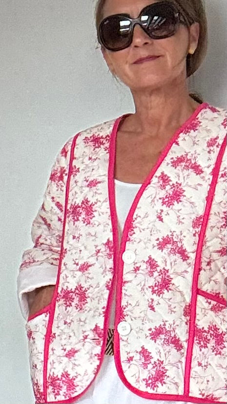 Steppjacke aus 100% Baumwolle in strahlendem Weiß mit pinkem Blumenmuster. Die Jacke kann offen oder mit eleganten Knöpfen geschlossen getragen werden. Mit einem oversized Schnitt und einer Rückenlänge von 56 cm passt sie perfekt über weite Kleider oder Röcke. Besondere Details sind aufgesetzte Taschen und feine Ziernähte. Ideal für kühlere Tage und bietet hohen Tragekomfort.