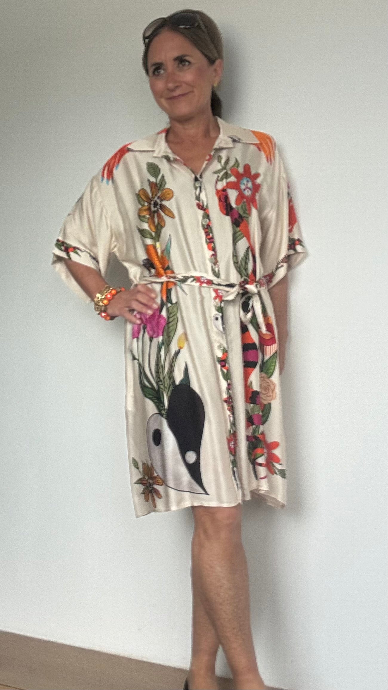 Blusenkleid aus 100 % Viskose mit ecrufarbenem Grundton und floralem Muster. Länge ca. 91 cm, locker geschnitten, mit Knopfleiste und Bindegürtel in der Taille. Auf der Rückseite des Kleides ist eine Göttin auf einem Leopard abgebildet – ein auffälliges, mystisch inspiriertes Detail. Das Kleid wird einmal geschlossen als Kleid und einmal offen als Kaftan über einer Hose gezeigt
