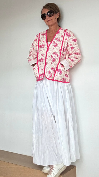 Steppjacke aus 100% Baumwolle in strahlendem Weiß mit pinkem Blumenmuster. Die Jacke kann offen oder mit eleganten Knöpfen geschlossen getragen werden. Mit einem oversized Schnitt und einer Rückenlänge von 56 cm passt sie perfekt über weite Kleider oder Röcke. Besondere Details sind aufgesetzte Taschen und feine Ziernähte. Ideal für kühlere Tage und bietet hohen Tragekomfort.