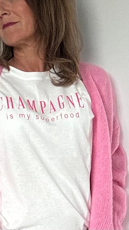 Cremeweißes T-Shirt mit einem lockeren Halsausschnitt und kurzen Ärmeln, auf der Brust steht in Pink geschreiben: Champagne is my superfood. Details findest du in unserer Produktbeschreibung.
