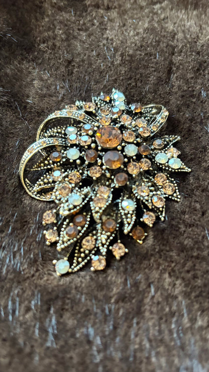 Oval-längliche Brosche mit bernsteinfarbenen, champagnerfarbenen und opalfarbenen Kristallsteinen auf goldener Metallfassung, fotografiert auf braunem Faux-Fur.