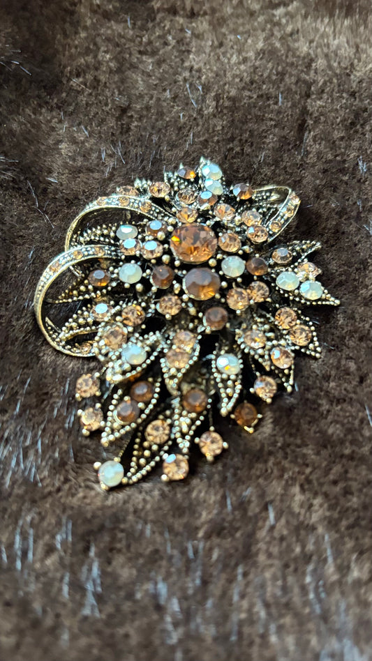 Oval-längliche Brosche mit bernsteinfarbenen, champagnerfarbenen und opalfarbenen Kristallsteinen auf goldener Metallfassung, fotografiert auf braunem Faux-Fur.