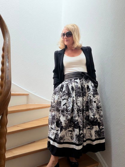 Frau trägt voluminösen Toile de Jouy Rock in Schwarz-Weiß mit schlichtem cremefarbenem Top, schwarzem Blazer und breitem Gürtel auf einer Holztreppe. Der Look wirkt modern, feminin und couture-inspiriert mit französischer Eleganz.
