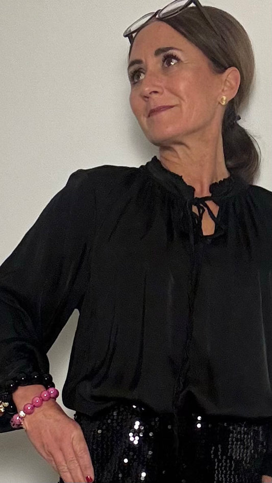 Leichte, elegante Bluse 'Ruffle Grace' aus 100% Viskose, leicht glänzend. Die Bluse hat einen lockeren, geraden Schnitt, einen kleinen Stehkragen mit Rüschen, lange Ärmel mit Knopfverschluss und eine Bindeschleife vorne. Perfekt kombinierbar mit Jeans, Röcken oder unter Strickjacken und Blazern. Länge 63 cm, Achsel zu Achsel 55 cm. Passt von Größe 34 bis schmaler 40, abhängig von der Oberweite.