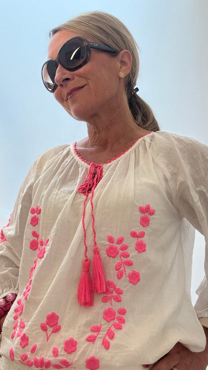 Weiße Boho-Bluse aus Baumwolle mit pinker floraler Stickerei auf Vorderseite und Ärmeln. Lockere Passform mit 3/4-Ärmeln und Bindebändern am Ausschnitt. Luftig, feminin und ideal für den Sommer, kombiniert mit dunkler Jeans.