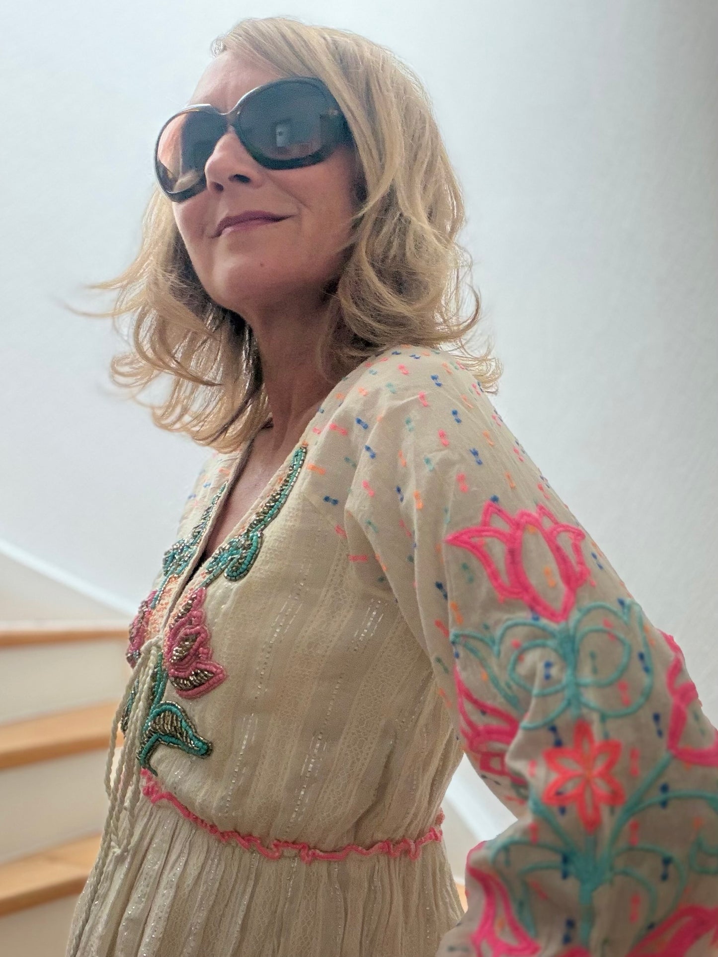 Frau trägt cremefarbenes Boho-Maxikleid aus Baumwolle mit pink-blauer Blütenstickerei, feinen Glanzfäden, V-Ausschnitt mit Troddeln und langen bestickten Ärmeln auf einer Holztreppe. Das Kleid wirkt leicht, feminin und sommerlich elegant.
