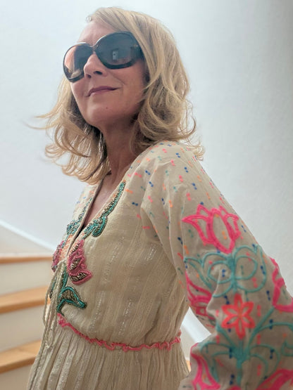 Frau trägt cremefarbenes Boho-Maxikleid aus Baumwolle mit pink-blauer Blütenstickerei, feinen Glanzfäden, V-Ausschnitt mit Troddeln und langen bestickten Ärmeln auf einer Holztreppe. Das Kleid wirkt leicht, feminin und sommerlich elegant.
