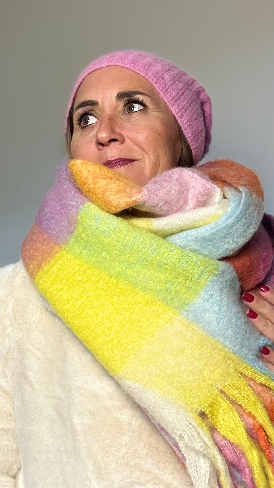 Langer, weicher Schal mit breiten Fransen und leuchtenden Farben in Rosa, Gelb, Hellblau und Orange. Der Schal liegt locker über einem hellen Mantel und wirkt farbenfroh und kuschelig.