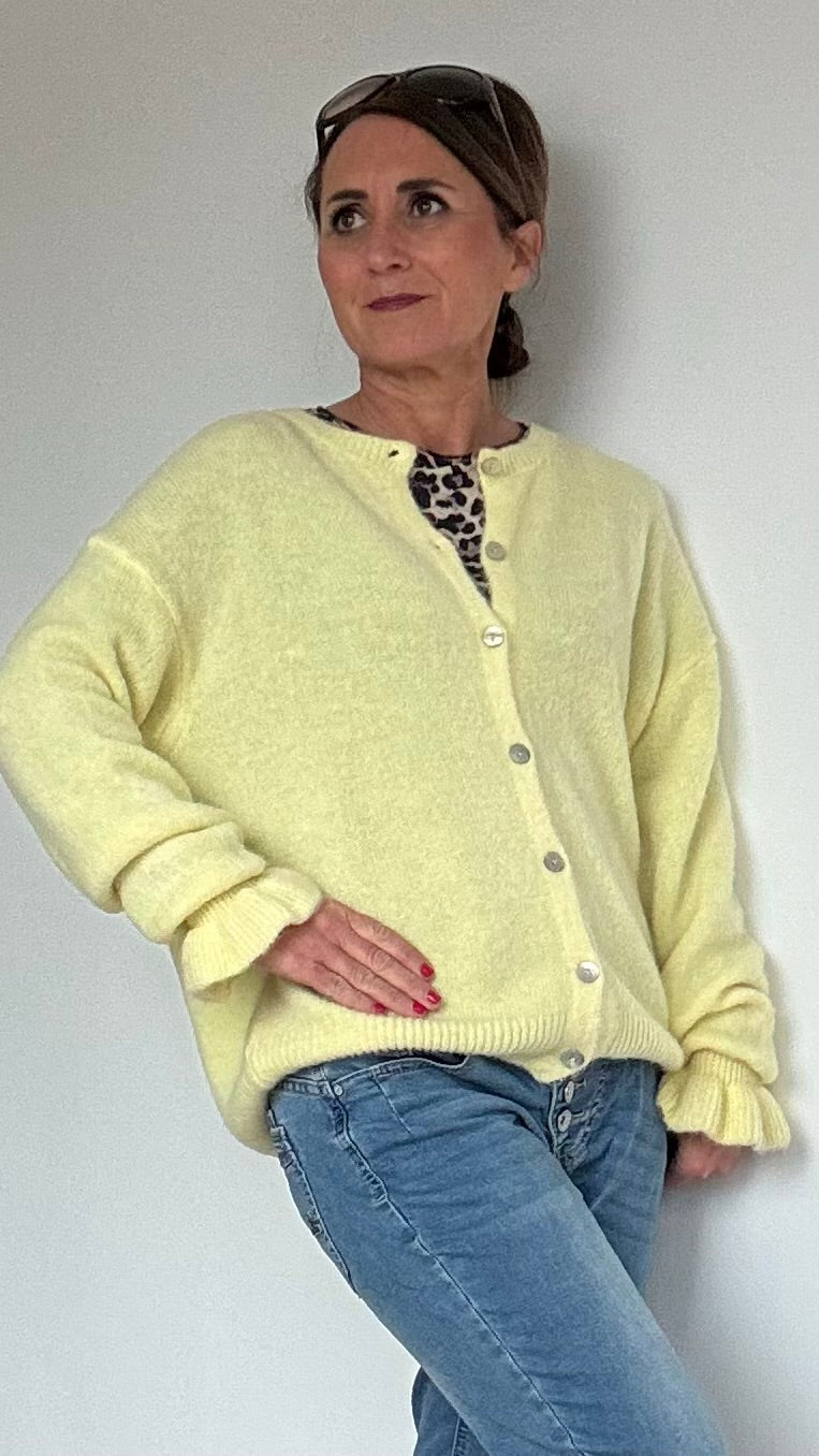 Ruffle Grace Cardigan in der Farbvariante hellgelb und lockerer Passform mit einer Länge von ca. 58 cm. Gefertigt aus 30 % Alpaka, 30 % Polyamid, 20 % Polyacryl, 17 % Wolle und 3 % Elastan. Der Cardigan hat einen Rundhalsausschnitt, lange Ärmel mit besonderen Strick-Rüschen an den Bündchen und Perlmuttknöpfe. Geeignet für Größen 34-42, ideal für feminine und vielseitige Looks. Kombinierbar mit Jeans, Röcken oder Kleidern für elegante und lässige Outfits