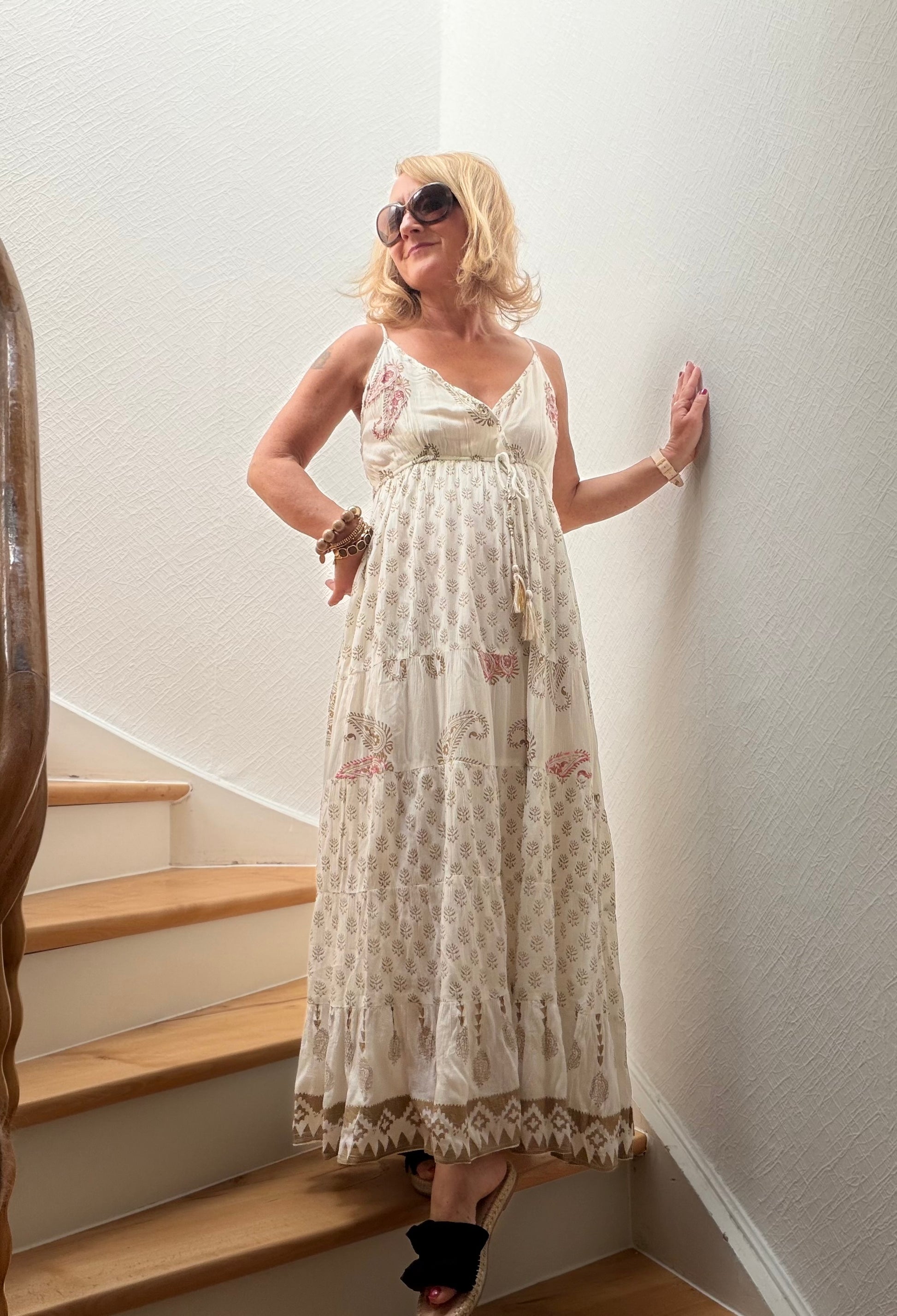 Frau trägt langes Boho-Maxikleid aus fließender Viskose mit feinen Naturmustern, Goldakzenten, gekreuztem V-Ausschnitt und verstellbaren Trägern auf einer Holztreppe. Das Kleid wirkt leicht, feminin und sommerlich elegant.
