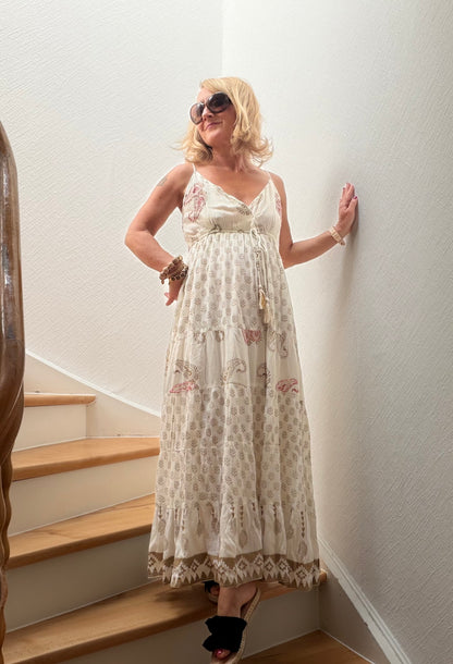 Frau trägt langes Boho-Maxikleid aus fließender Viskose mit feinen Naturmustern, Goldakzenten, gekreuztem V-Ausschnitt und verstellbaren Trägern auf einer Holztreppe. Das Kleid wirkt leicht, feminin und sommerlich elegant.
