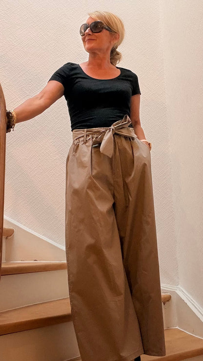 Miss Lotti trägt eine taupefarbene weit geschnittene Marlenehose mit fließendem Bein, elastischem Bund und Bindegürtel. Dazu ein enges T-Shirt in Schwarz.
