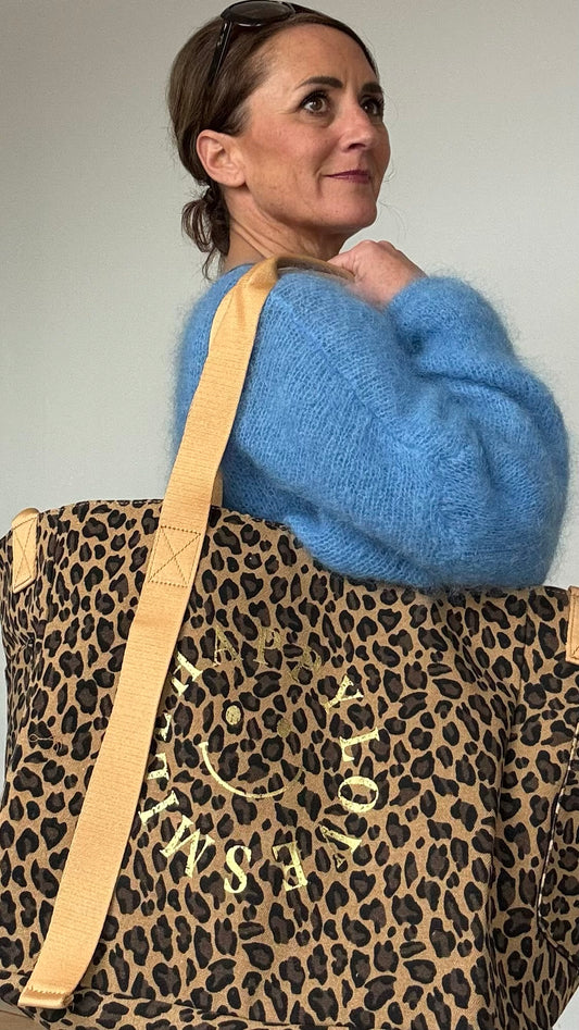 Großer Maxi-Shopper aus robustem Canvas im dunklen Leoprint mit einem goldenen Print in Form eines Smiley ohne runden Rand. Die Worte 'Happy Smile Love' formen den Umriss des Smiley-Designs. Die Tasche misst 35 cm in der Höhe, 40 cm in der Länge und 28 cm in der Breite. Innen ist die Tasche schwarz gefüttert und hat eine große Innentasche mit Reißverschluss. Zwei lange Tragegurte ermöglichen das Tragen über der Schulter, während zwei kleinere Henkel an den Seiten ein bequemes Tragen in der Hand ermöglichen.