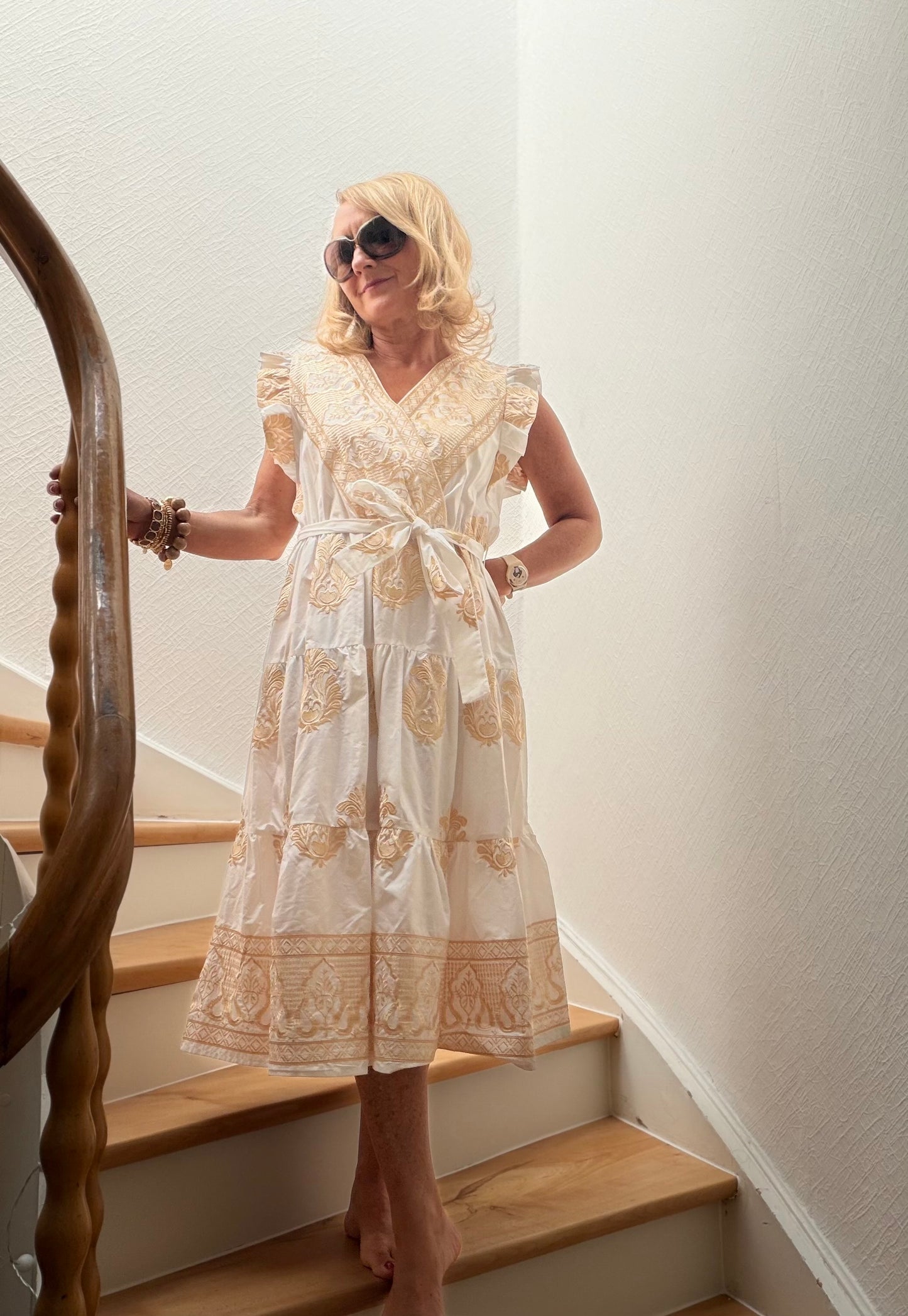 Frau trägt cremefarbenes Midikleid aus Baumwolle mit goldener Stickerei, überkreuztem Oberteil, kleinen Flügelärmeln und femininer A-Linie auf einer Holztreppe. Das Kleid wirkt leicht, elegant und sommerlich und eignet sich besonders gut für Petite-Frauen.
