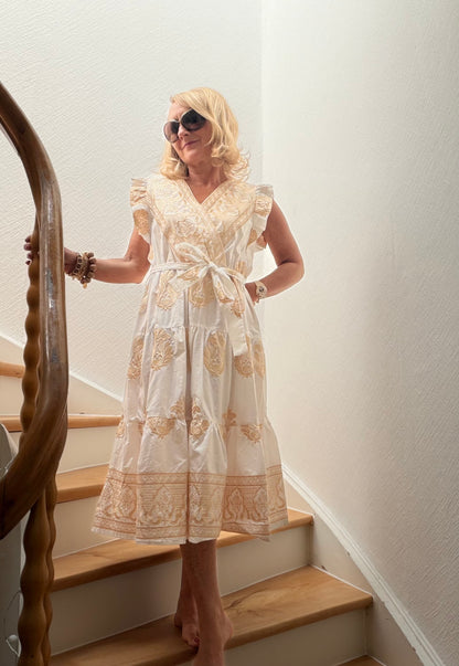 Frau trägt cremefarbenes Midikleid aus Baumwolle mit goldener Stickerei, überkreuztem Oberteil, kleinen Flügelärmeln und femininer A-Linie auf einer Holztreppe. Das Kleid wirkt leicht, elegant und sommerlich und eignet sich besonders gut für Petite-Frauen.
