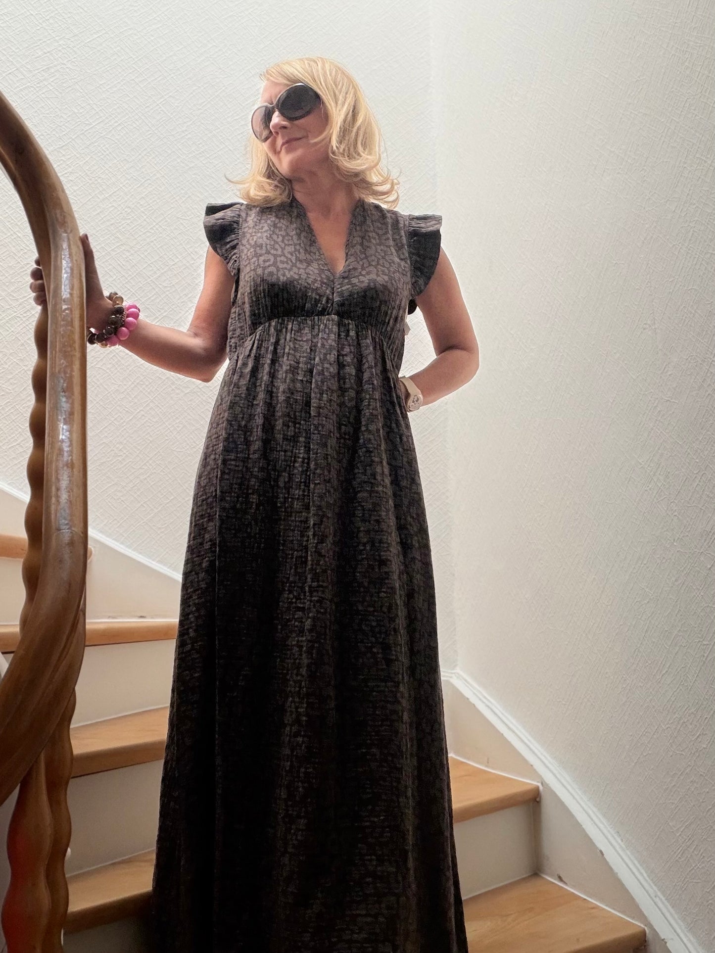 Frau trägt langes Musselin-Maxikleid aus dunkelbrauner Baumwolle mit taupefarbenem Leo-Print, V-Ausschnitt und kleinen Flügelärmeln auf einer Holztreppe. Das Kleid wirkt leicht, feminin und sommerlich entspannt.
