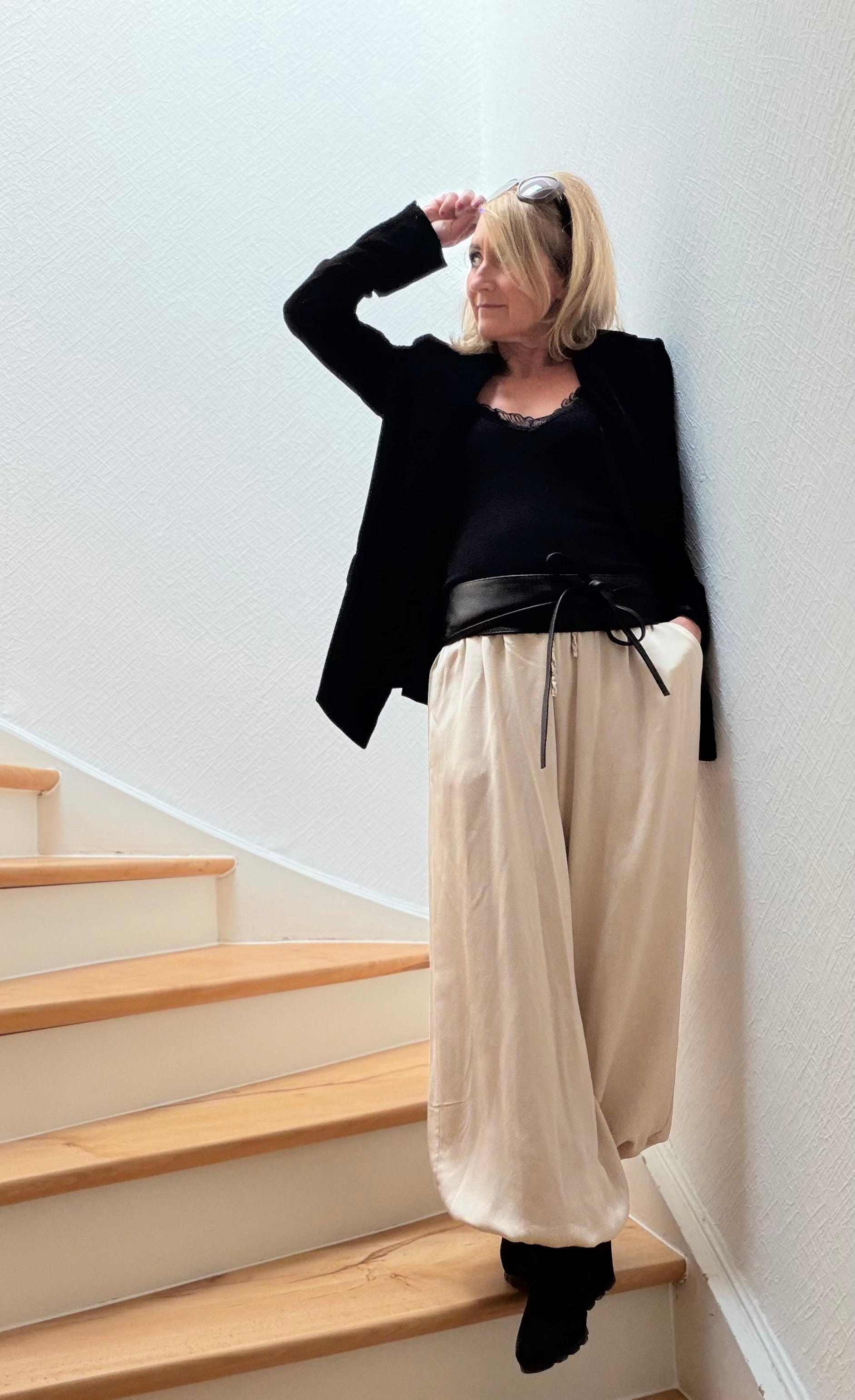 Petite Frau trägt eine fließende Pluderhose aus Viskose-Seide in Beige mit elastischem Bund und Bündchen am Saum. Die Hose wird mit einem schwarzen Spitzentop kombiniert und locker auf einer Treppe gestylt. Weicher Fall, leicht glänzende Oberfläche und figurschmeichelnde Silhouette durch betonte Taille.