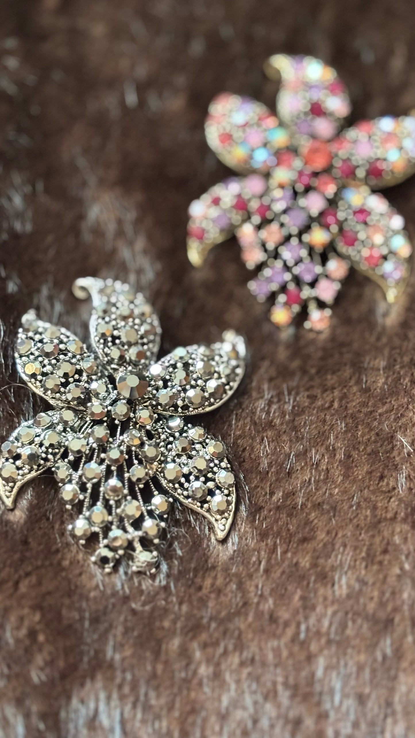 wei florale Kristallbroschen in Blütenform: eine in Silber-Champagne, eine in Rosé-Multicolor. Beide liegen auf braunem Faux-Fur und zeigen ihre funkelnden Details