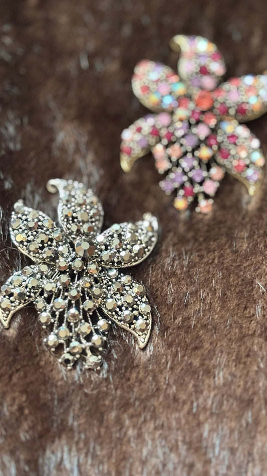 wei florale Kristallbroschen in Blütenform: eine in Silber-Champagne, eine in Rosé-Multicolor. Beide liegen auf braunem Faux-Fur und zeigen ihre funkelnden Details