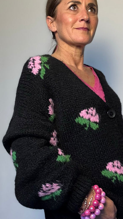 Kuschelig weicher Strickcardigan mit V-Ausschnitt, Knopfleiste und romantischen eingestrickten Blumen in Rosa. Ballonärmel mit schmalem Bündchen. Erhältlich in Schwarz und Wollweiß, passt von Größe 34 bis 40. Perfekt für stilvolle Layering-Looks in der Übergangszeit.