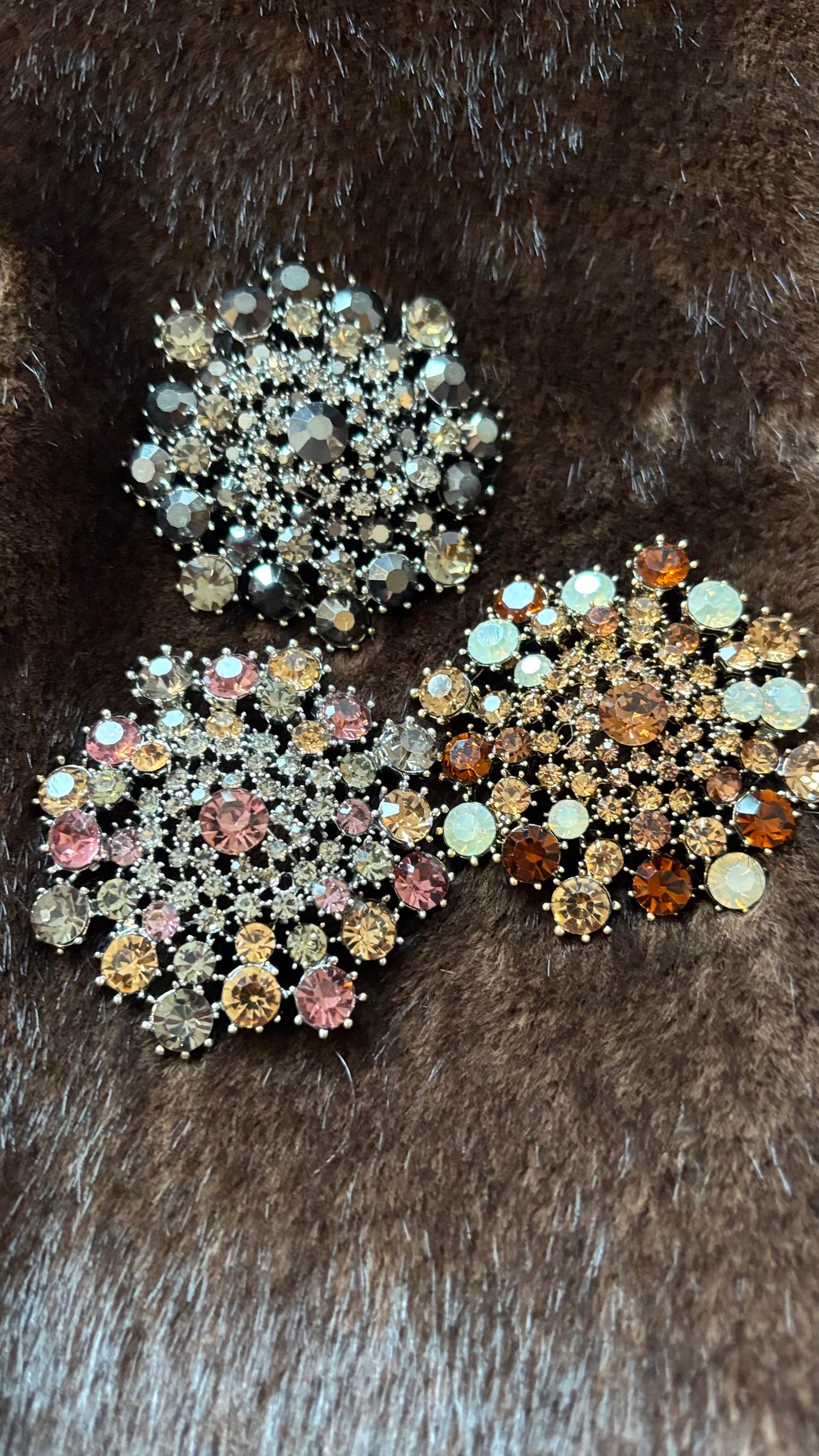 Drei runde, ca. 5,5 cm große Broschen mit funkelnden Kristallsteinen, in den Farbkombinationen Rosé, Amber und Black-Champagne. Die Broschen liegen auf braunem Faux-Fur-Hintergrund und zeigen ihre detailreichen Steinsetzungen und warmen Metallfassungen.