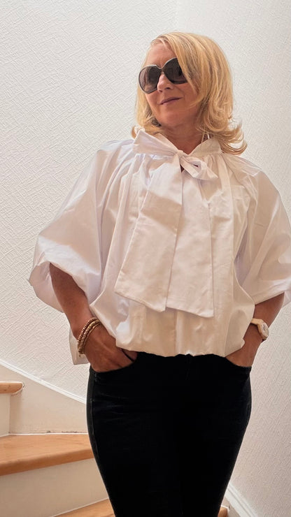Miss Lotti  trägt eine weiße Ballonbluse mit weiten 3/4-Ärmeln, gerafftem Saum und Bindeschleife am kleinen Stehkragen.